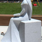 A side view of a granite sculpture depicting a seated woman in a flowing gown holding an open book on her lap. The setting includes a green lawn and construction equipment in the background.