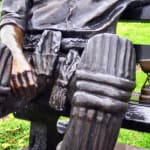 Detail of Keith Miller’s bronze statue, focusing on the protective cricket pads and a miniature Ashes urn placed beside him on the bench, symbolizing his significant contribution to the sport.