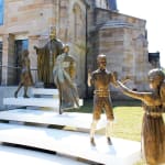 A bronze sculpture installation outside a stone church, featuring Pope John Paul II blessing a group of figures. The composition includes a young man draped in an Australian flag, a young woman approaching the Pope, and two children joyfully interacting in the foreground. The sculptures are mounted on tiered white platforms with engraved text, set against the backdrop of the church's sandstone facade and stained glass doors. The scene conveys themes of faith, community, and youthful energy.