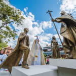 A bronze sculpture installation featuring multiple life-sized figures, including a young man stepping forward with an Australian Flag over his shoulder, a young woman standing attentively, and a bishop holding a pastoral staff. The Archbishop of Sydney in ceremonial robes blesses the statues with holy water as acolytes and onlookers stand nearby. The scene is set outdoors on a sunny day, with a tree and historic buildings in the background.