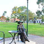 A full view of the bronze statue of Mayor Joy Baluch, seated in a chair next to a table with a rose placed on the base of the statue. The setting is an open park with trees, pathways, and lampposts, emphasizing the public nature of the memorial.