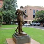 The back view of the Shore Boys bronze statue shows the man walking forward with his arm around the boy, symbolizing guidance and support, set against the lush green school grounds.