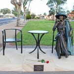 The bronze statue of Joy Baluch, seated next to a chair and table, on a sandstone base inscribed with plaques. The setting includes a park pathway and a rose placed in front of the table, honoring the late mayor's legacy in Port Augusta.