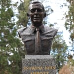 A bronze bust sculpture of Anthony John Abbott, former Prime Minister of Australia, mounted on a polished granite pedestal. The bust depicts Abbott wearing a shirt and tie, with a slight smile and finely detailed facial features. The pedestal is inscribed with gold lettering reading, "ANTHONY JOHN ABBOTT / PRIME MINISTER / 2013 — 2015." The setting is an outdoor environment with lush green trees and a white fence in the background, emphasizing the dignified and commemorative nature of the sculpture.