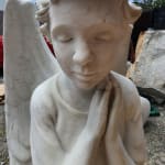 A close-up view of a marble statue depicting a serene angel with closed eyes and clasped hands in prayer. The angel's soft features and detailed curls convey peace and spirituality, set against an urban background with modern architecture.