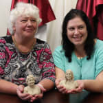 A candid moment showing a mother and daughter holding mini-bust sculptures modeled in plastilina. The mother, with curly white hair, wears a patterned red and black outfit, while the daughter, with long dark hair, smiles in a mint green blouse. Each holds a likeness of themselves, highlighting their connection to the artistic process.