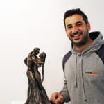 Andrew Christou, smilingLovers bronze statuette that he commissioned as a wedding gift for his wife. The sculpture depicts a couple in an embrace, symbolizing love and devotion, showcased on a white pedestal.