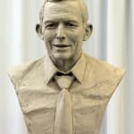 A clay bust sculpture of Anthony John Abbott, former Prime Minister of Australia, in progress. The bust features detailed facial expressions and a textured surface, showing Abbott in a collared shirt and tie. The unfinished texture highlights the sculpting process, emphasizing the artist's craftsmanship. The background is neutral with soft, white draping, drawing attention to the clay model.