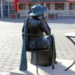 The back view of the statue of Mayor Joy Baluch, showing her seated in the chair with a wide-brimmed hat and a shawl draped over her shoulder. The background includes a historic building and street with visible signs of the Port Augusta community.
