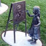 Bronze sculpture of a little girl smiling and painting on an easel, set in a garden environment.