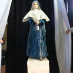 A resin statue of Jeanne-Marie Chavoin, founder of the Marist Sisters, unveiled against a black curtain backdrop. The statue captures Jeanne-Marie in her traditional religious habit, including a flowing blue gown and a white veil. She holds a cross in one hand and a lantern in the other, symbolizing faith and guidance. The sculpture stands on a white marble-like pedestal, with a warm, welcoming expression on her face. The unveiling is marked by curtains being pulled aside on either side, adding a ceremonial atmosphere to the presentation.