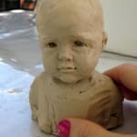 A miniature bust of a child in plastiline, showcasing a soft and innocent expression. The sculpture captures the delicate features of a young face, with subtle details in the hair and garment folds.