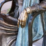 A detailed close-up of the statue, highlighting Joy Baluch's hand resting on the armrest, adorned with bracelets, and the textured folds of her dress and shawl. The artistry of the bronze details is emphasized.