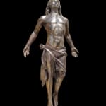 Bronze statue of Jesus ascending, depicted with outstretched arms and upward gaze. His muscular figure is draped with a flowing cloth around his waist, conveying movement and spiritual elevation. The background is white, emphasizing the sculpture's detailed craftsmanship.