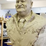 An angled view of the clay bust of Lindsay Fox, showcasing his distinctive facial features and the unfinished, textured surface of the sculpture. The figure is dressed in a jumper with a collared shirt and tie, placed in a workshop setting.