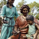 A bronze statue of the Holy Family, showing Mary, Joseph, and the child Jesus in an intimate and joyful moment. Joseph is seated, holding the child on his lap, while guiding the child’s hands toward a dove. Mary stands beside them, smiling warmly, with her hand resting on Joseph’s shoulder.
