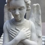 A close-up of a marble angel statue with closed eyes and crossed hands over the chest. The intricate details of the wings and the delicate curls in the hair emphasize the craftsmanship of this contemplative figure.