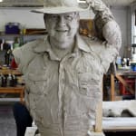 Close-up of a clay bust of Lord Alistair McAlpine in progress. He is wearing a wide-brimmed hat and a textured button-up shirt. A detailed black cockatoo perches on the brim of the hat, adding a unique element to the sculpture. The bust captures a warm and smiling expression