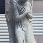 A full view of a kneeling marble angel statue with intricately carved wings, hands crossed over its chest, and a flowing robe. The statue captures a moment of quiet devotion, standing against a plain backdrop.