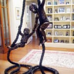 A bronze sculpture of two young boys playing on ropes, displayed indoors on a polished wooden surface. One boy climbs the rope confidently while the other, slightly younger, joyfully swings from a looped section, their expressions capturing a playful and adventurous spirit. Behind the sculpture is a bookshelf filled with framed family photographs, adding a warm, personal backdrop to the scene.