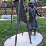 Bronze sculpture of a little girl smiling and painting on an easel, set in a garden environment.