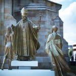 A bronze sculpture installation featuring multiple life-sized figures, including a young man stepping forward with an Australian Flag over his shoulder, a young woman standing attentively, and St John Paul II holding a staff topped with a crucifix. The scene is set outdoors on a sunny day, with the sandstone wall of St Patrick's Cathedral in the background.