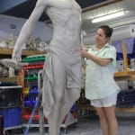 Artist Linda Klarfeld sculpting a life-size clay statue of Jesus ascending. The sculpture features Jesus with outstretched arms and an upward gaze, draped in a flowing cloth around his waist. The scene is set in a studio filled with sculpting tools and materials.