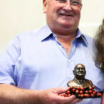 Lindsay Fox, wearing glasses and a light blue shirt, smiles as he holds a miniature bronze bust of himself. The bust is mounted on a base surrounded by bright red Jaffa lollies, symbolizing his personality: hard on the outside but soft on the inside.