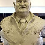 A clay model of a bust of Lindsay Fox, capturing his strong features and dressed in a jumper with a collared shirt and tie. The textured clay gives a raw and unfinished appearance, emphasizing the sculpting process.