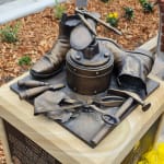 Another perspective of the bronze still-life memorial for Jim Saad, emphasizing the intricate details of the cobbler's tools and a boot, with flowers visible at the base, highlighting the community's tribute to his legacy.