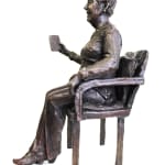 A bronze statuette of a seated woman, named Audrey, holding a book in her left hand. She is depicted wearing detailed clothing with intricate textures and patterns. The figure is seated on a chair with armrests, exuding a sense of warmth and personality.