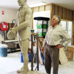 Neil Roberts, a celebrated Australian Rules footballer, is seen as an older man standing beside the clay model of his younger self. The clay statue captures him in his prime, wearing his playing uniform and holding a football with both hands. Roberts observes the artwork closely, his admiration evident as the artist works to perfect the fine details, showcasing his athletic physique and determined expression.