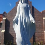 A granite statue of Mary, titled 'Mary, Star of the Sea,' standing gracefully in front of a brick church building under a bright blue sky. The statue features Mary with outstretched arms, flowing robes, and a serene expression, evoking peace and reverence. The church’s tiled roof and a cross at the peak of the structure create a harmonious background for the sculpture.