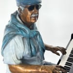 Detail view of upper torso and hands of the bronze sculpture of an elderly man seated at a grand piano, playing with a serene and focused expression. The figure is dressed casually in shorts, a t-shirt, and a sweater draped over his shoulders, with vibrant color accents highlighting his clothing and shoes. The artwork captures a moment of musical engagement and nostalgia in a modern indoor setting.