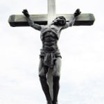 ronze sculpture of Jesus on the cross, shown from the front view. The detailed musculature and expression capture the solemn moment of crucifixion, with the cross standing against a pale sky.
