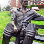 Bronze statue of Keith Miller seated on a bench, wearing cricket gear and exuding a relaxed and charismatic presence. The sculpture celebrates his stature as one of Australia’s cricketing legends, set against the backdrop of a historical school building.