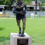 Bronze statue of rugby league legend Peter 'Pedro' Gallagher, depicted in a dynamic running pose while holding a rugby ball. The statue, mounted on a pedestal with a commemorative plaque, showcases intricate detailing of his uniform and captures the strength and determination of his character. The first image situates the sculpture in an outdoor park setting