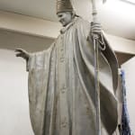 A clay model of a statue depicting Pope John Paul II in progress. The figure is shown wearing papal robes and a mitre, holding a staff topped with a crucifix in one hand and extending the other in a gesture of blessing. The sculpture is mounted on a platform covered with a blue tarp, inside a workshop with a plain white wall and fluorescent lighting.