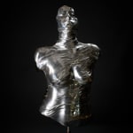 Frontal view of an aluminum torso sculpture, showing a polished, reflective surface with muscular detailing. The figure's head tilts upwards, suggesting a gesture of aspiration or contemplation, with smoky quartz crystals visible along the back.