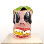 A large ceramic face jug by Kjelshus Collins. The face has green hair, six eyes, red lips, crooked teeth, and an ear of corn in its mouth