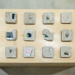 A sculpture by Shabez Jamal consisting of 16 polaroid photographs in various states of emergence from poured concrete squares.