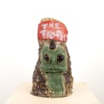 A ceramic pitcher by Kjelshus Collins. The front of the pitcher shows a green alien creature with a single horn. Above him a red wooden sign reads 'the tropics'