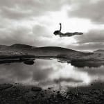 Silver gelatin print of Slate Gray Gallery artist Jerry Uelsmann's "Untitled, 1987 (Flying Figure Colorado) of a woman flying over a lake with mountains in the background in black and white