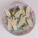 Ceramic sculpture of a bowl of bananas cut in half with faces and two peanuts with faces on a bed of lettuce