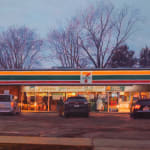 Painting of a 7/11 parking lot with three cars in the lot. There is a person going to the their car and another person dressed up as a ghost with a sheet over them