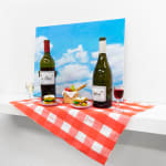 Installation featuring two bottles of wine with dog faces, two sandwiches with dog faces, a picnic basket with a baguette, an apple and a pear all with dog faces on top of a red and white checkered table clothe and a painted sky with clouds in the shape of dogs.