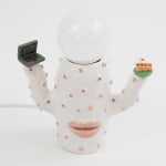 Jen Dwyer lamp sculpture of white cactus with pink lips holding laptop and slice of cake. Lightbulb sits on top