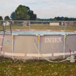 Painting of an above ground pool with the word "INTEX" in white on the side of the pool as well as a hose and a step ladder going into the pool. In the background is a chain fence and a few houses beyond the fence.