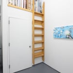 a white wall with a long blue and grey painting on the right, to its left a door and.a ladder leading to a shelf with paintings in storage and a window with a statue in it