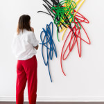 Scribble artwork from Arnaud Vidricaire made of laser cut wood with red blue green black and yellow overlapping colors with woman standing next to it for scale